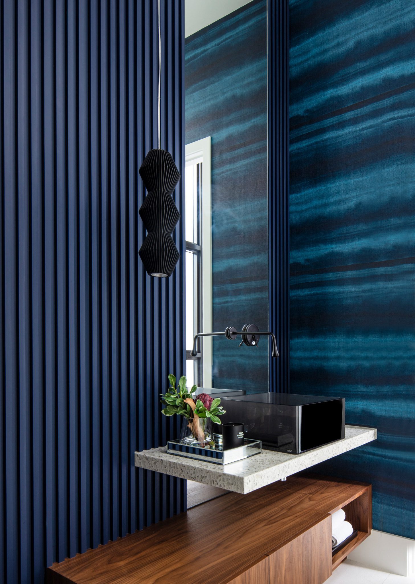 10-WEB-L Modern bathroom with dark blue walls, a floating sink with a rectangular mirror above, a black pendant light, and a wooden vanity below. A small tray with decorative items sits on the sink counter.