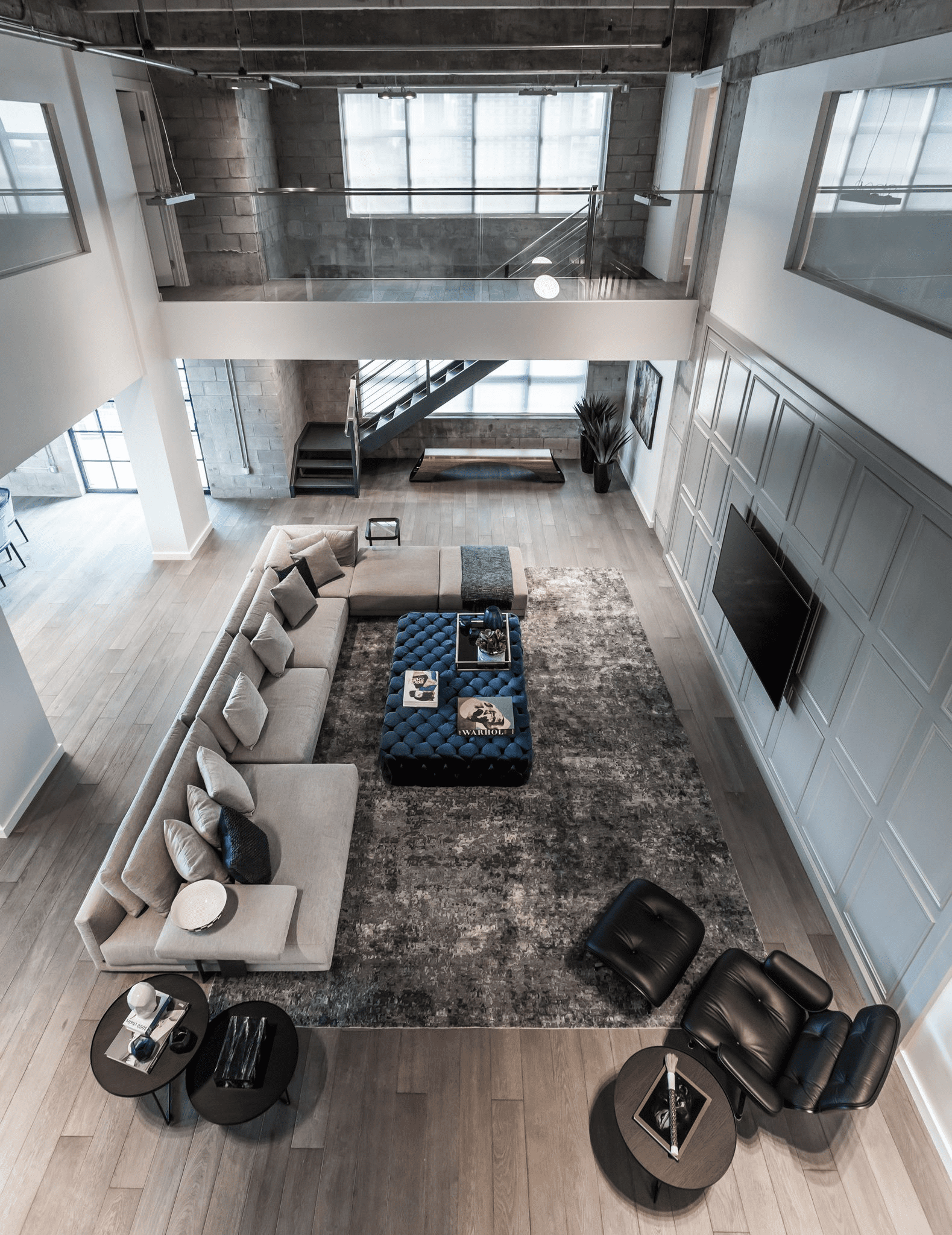 image (1) A modern, open-concept loft with high ceilings and large windows. It features a sectional sofa, a black recliner, and a coffee table on a gray rug in the living area. Stairs lead to an upper level.
