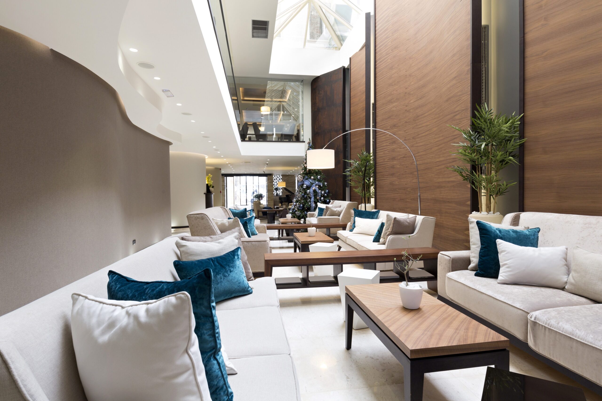 lobby1 A modern hotel lounge with white sofas, teal cushions, wooden tables, plants, and a high ceiling.